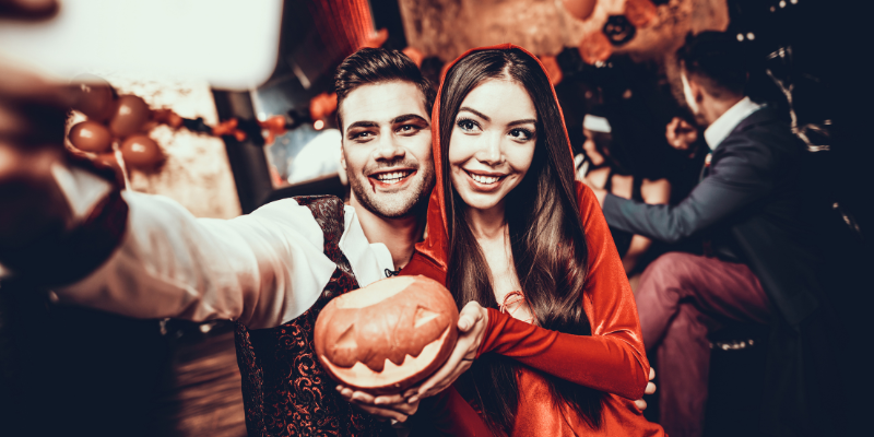 Two people dressed up in Halloween costumes taking a selfie with a phone. One is holding a pumpkin.
