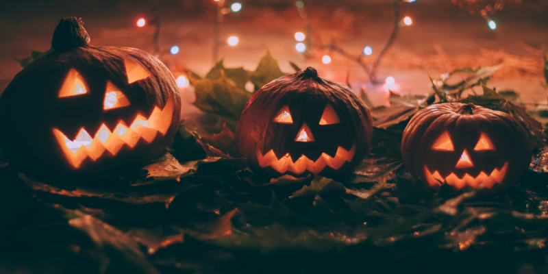 Three lit jack-o-lanterns sitting on a pile of leaves with string lights in the background