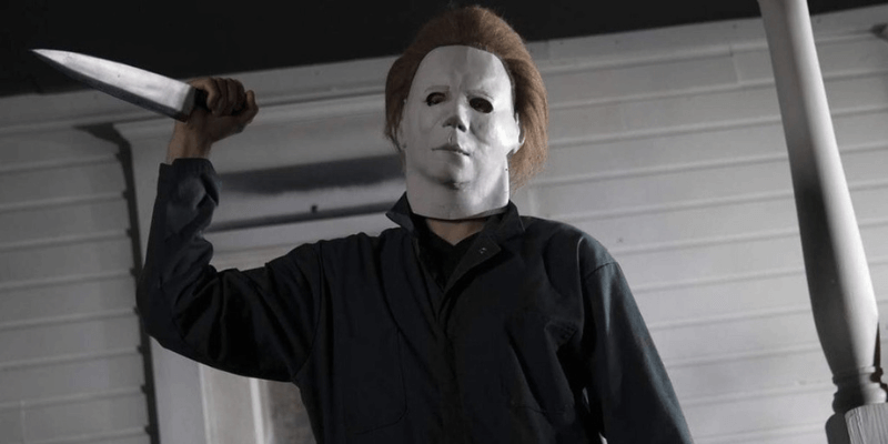 Michael Myers wearing his iconic white mask while standing in front of a house, holding up a large kitchen knife.