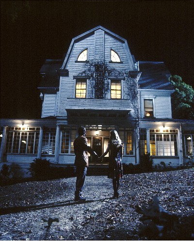 Two people standing outside in front of a dimly lit Amityville House at night.
